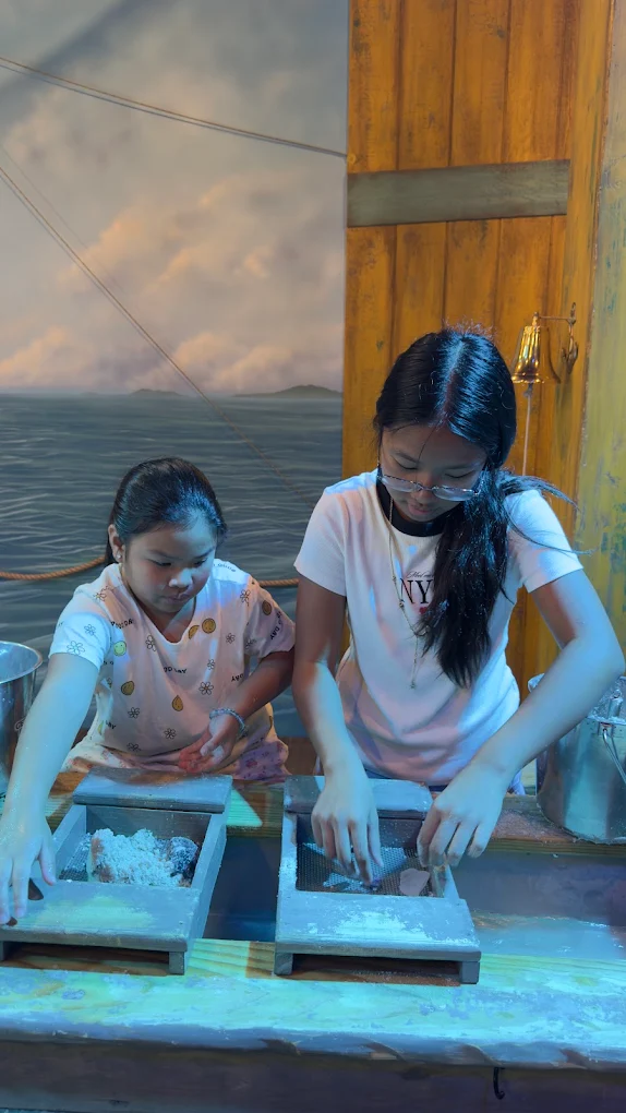 Two girls sifting for treasures at the mining trough in front of the painted sea backdrop